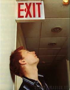 Thom apoyado en una pared con un cartel de exit arriba